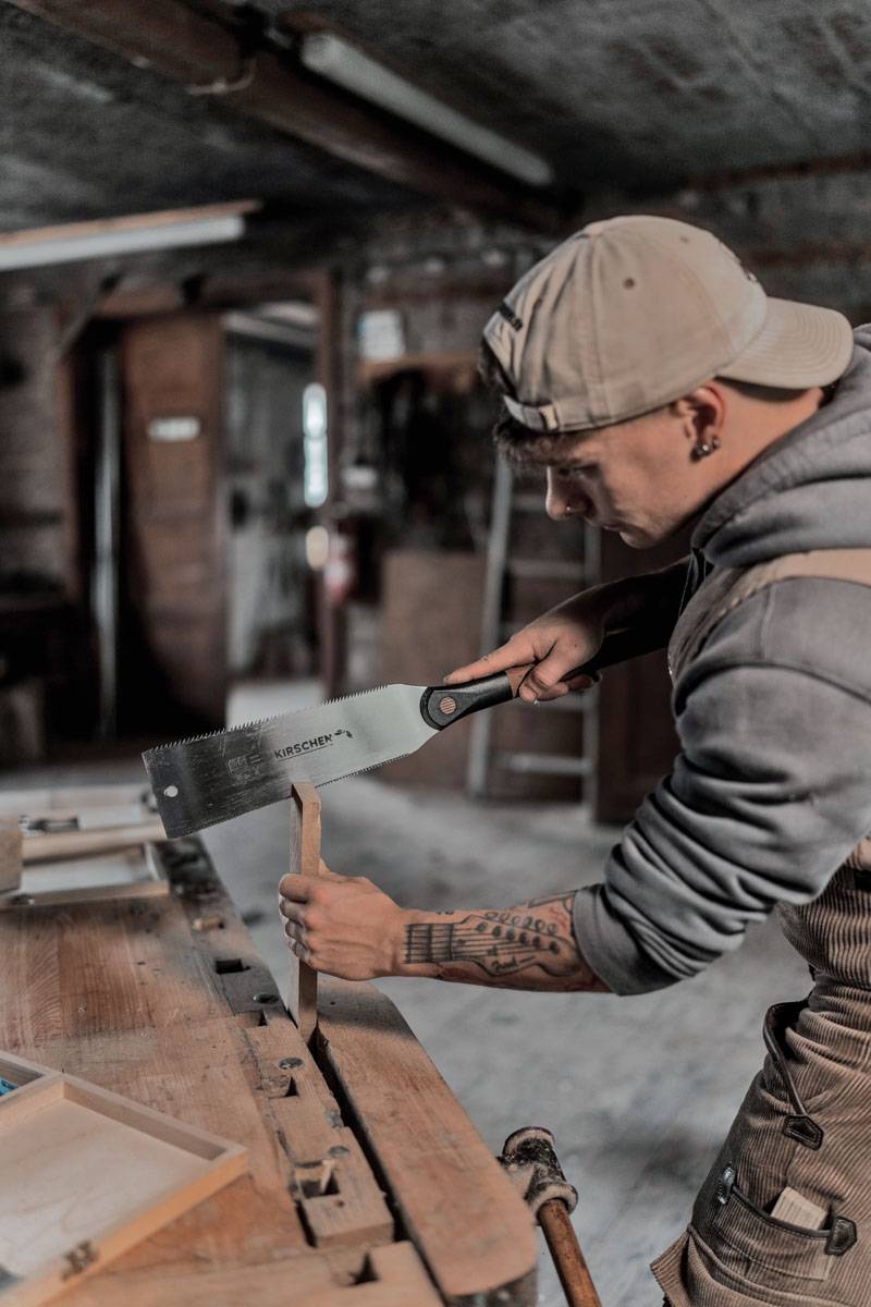 Eine Person, die eine Kappe und ein Sweatshirt trägt, benutzt eine Handsäge, um ein Stück Holz in einer Werkstatt zu schneiden. Der Fokus liegt auf der Sägebewegung.