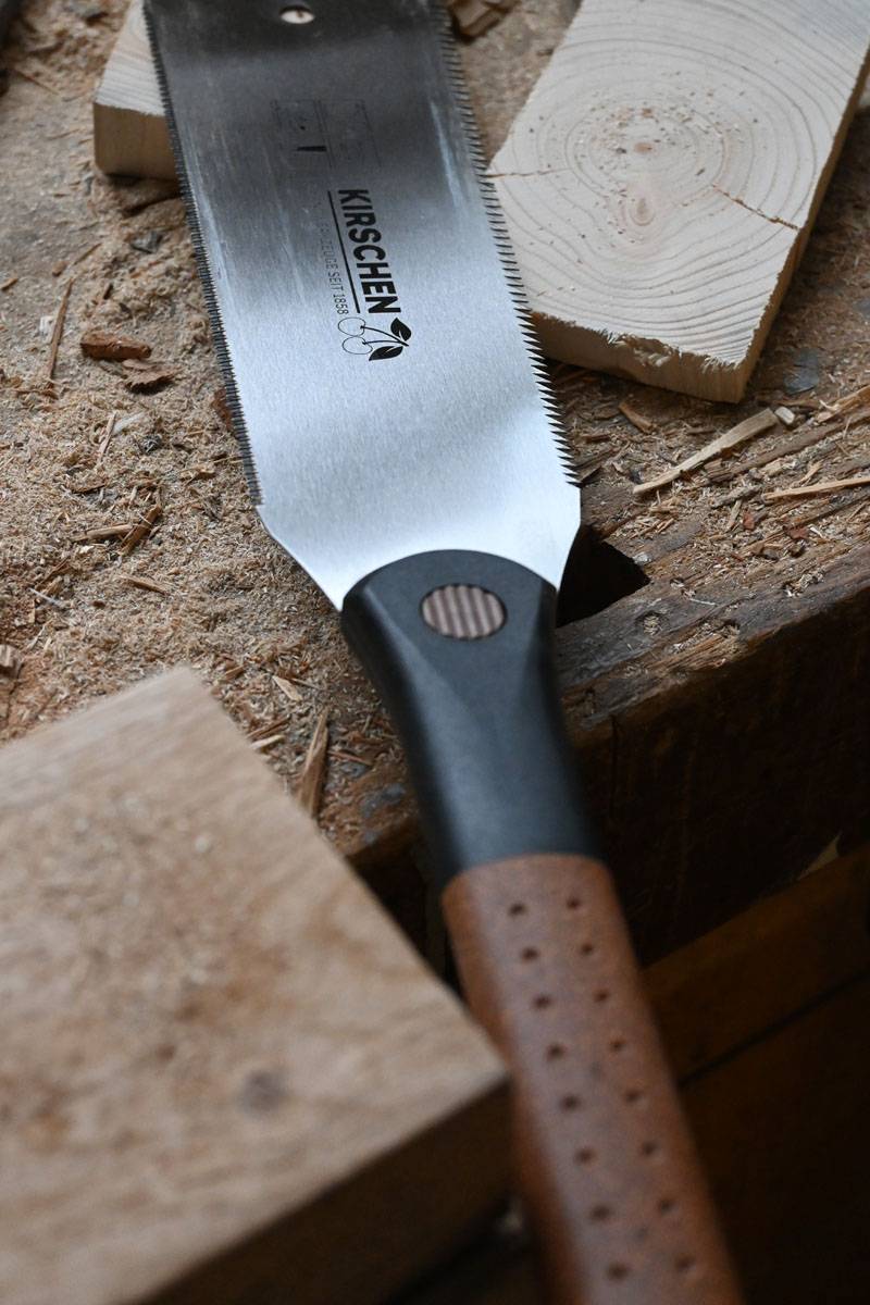 Eine Handsäge mit hölzernem Griff und scharfer, silberner Klinge ruht auf grob zugesägten Holzstücken auf einer Werkbank.