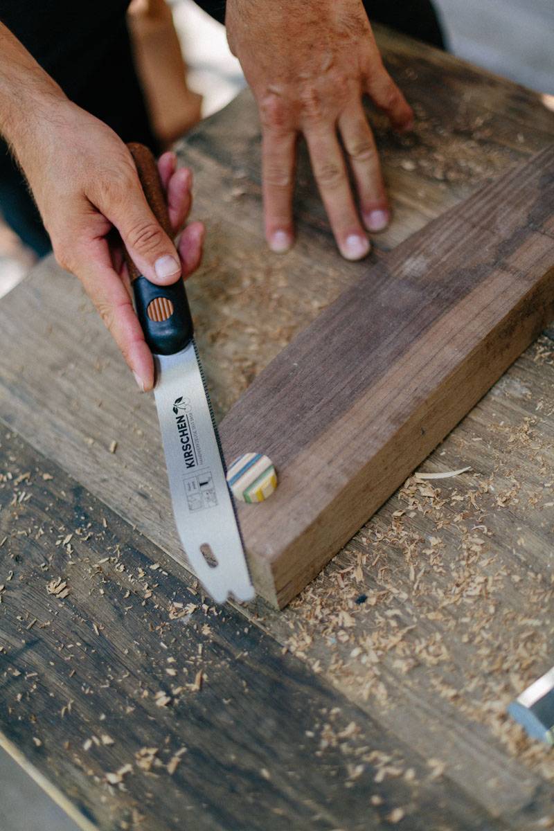 Eine Person nutzt eine Zugsäge, um einen Holzdübel bündig mit einer Holzoberfläche abzusägen, umgeben von Holzspänen. Die Hände zeigen Kontrolle und Präzision.