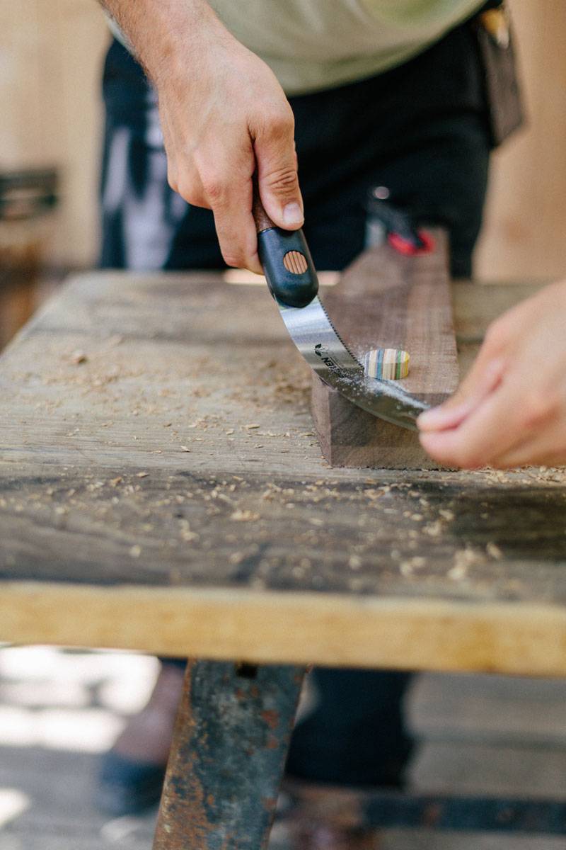 Eine Person benutzt einen Handhobel an einem Holzstück und glättet die Oberfläche. Sägespäne sind auf der Werkbank sichtbar.