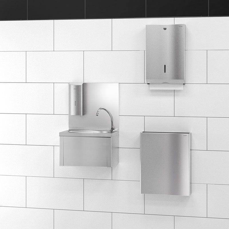 Drei an der Wand montierte Edelstahl-Badezimmerarmaturen auf weißem Fliesen: ein Waschbecken mit Wasserhahn, ein Spender und zwei geschlossene Einheiten.