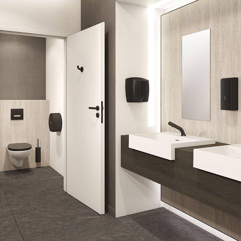 Modernes öffentliches Badezimmer mit zwei wandmontierten Toiletten auf der linken Seite und doppelten Waschbecken mit Spiegeln auf der rechten Seite; eine Tür steht in der Mitte offen.