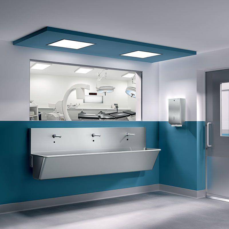 Krankenhausflur mit an der Wand montiertem Handwaschbecken außerhalb eines Fensters eines Operationssaals, das medizinische Ausrüstung im Operationssaal zeigt.