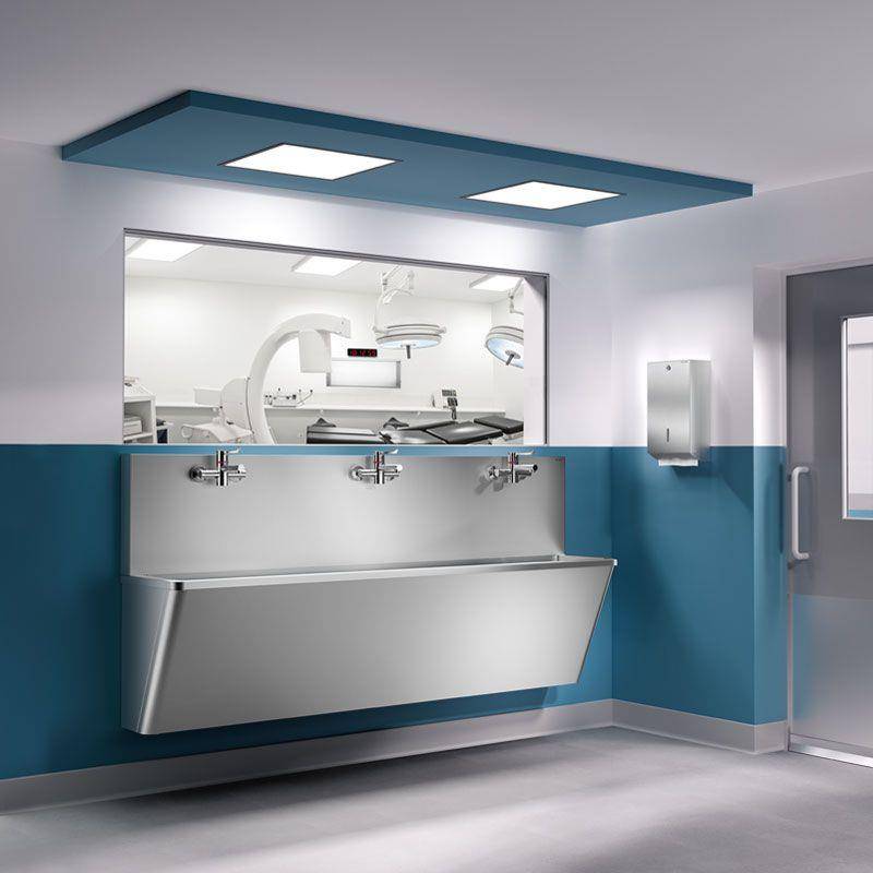 Steriles Krankenhausspülbecken mit drei Wasserhähnen in einem blau-weißen Flur neben einem Glasfenster in einen Operationssaal.