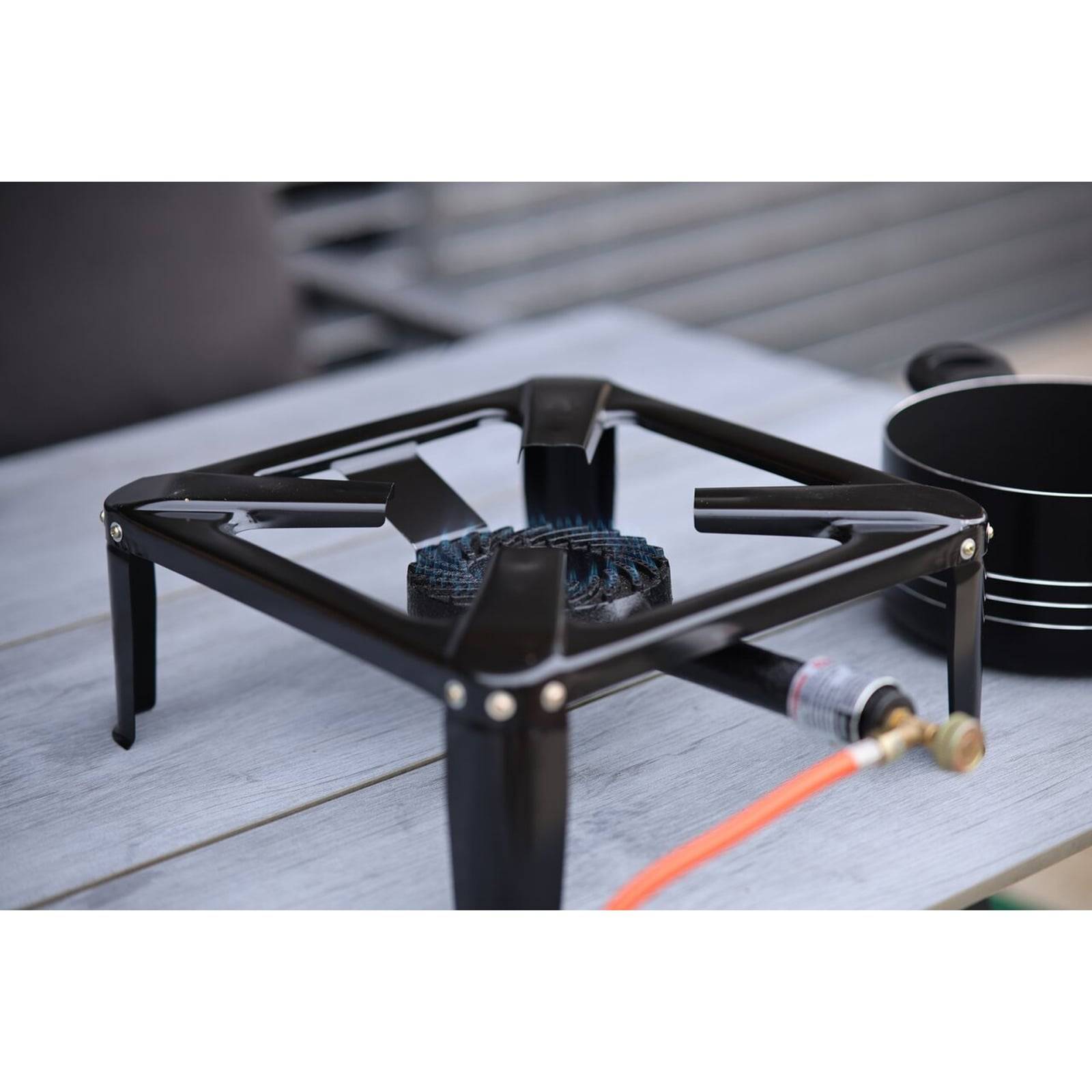 Ein tragbarer Gaskocher mit schwarzem Rahmen und Brenner auf einem Tisch im Freien, der über einen orangefarbenen Schlauch an einen Propantank angeschlossen ist. Daneben steht ein schwarzer Topf.