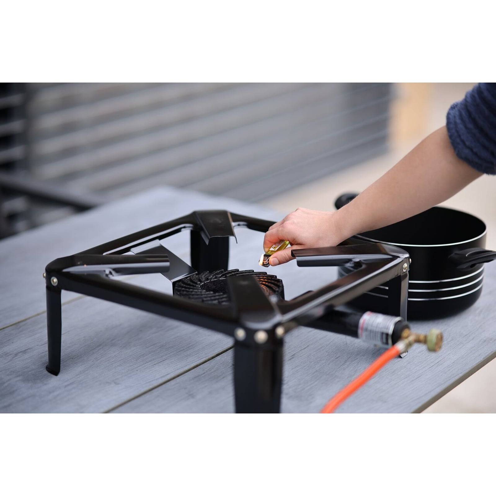 Eine Person zündet einen tragbaren Gaskocher auf einem Tisch an und bereitet sich auf das Kochen im Freien vor. Schwarze Töpfe sind daneben gestapelt.