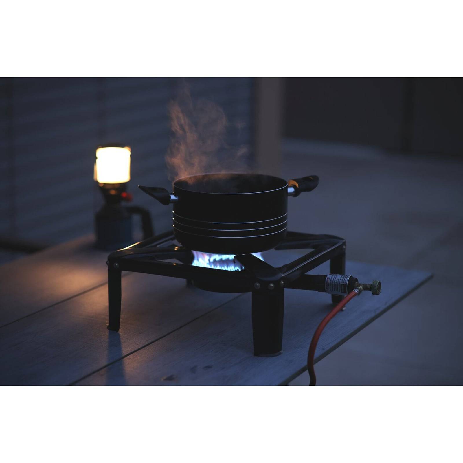 Ein Topf mit Dampf steigt auf einem tragbaren Gasherd auf, blauer Flammenschein ist sichtbar. Eine Laterne steht in der Nähe und deutet auf einen Outdoor-Kochplatz hin.