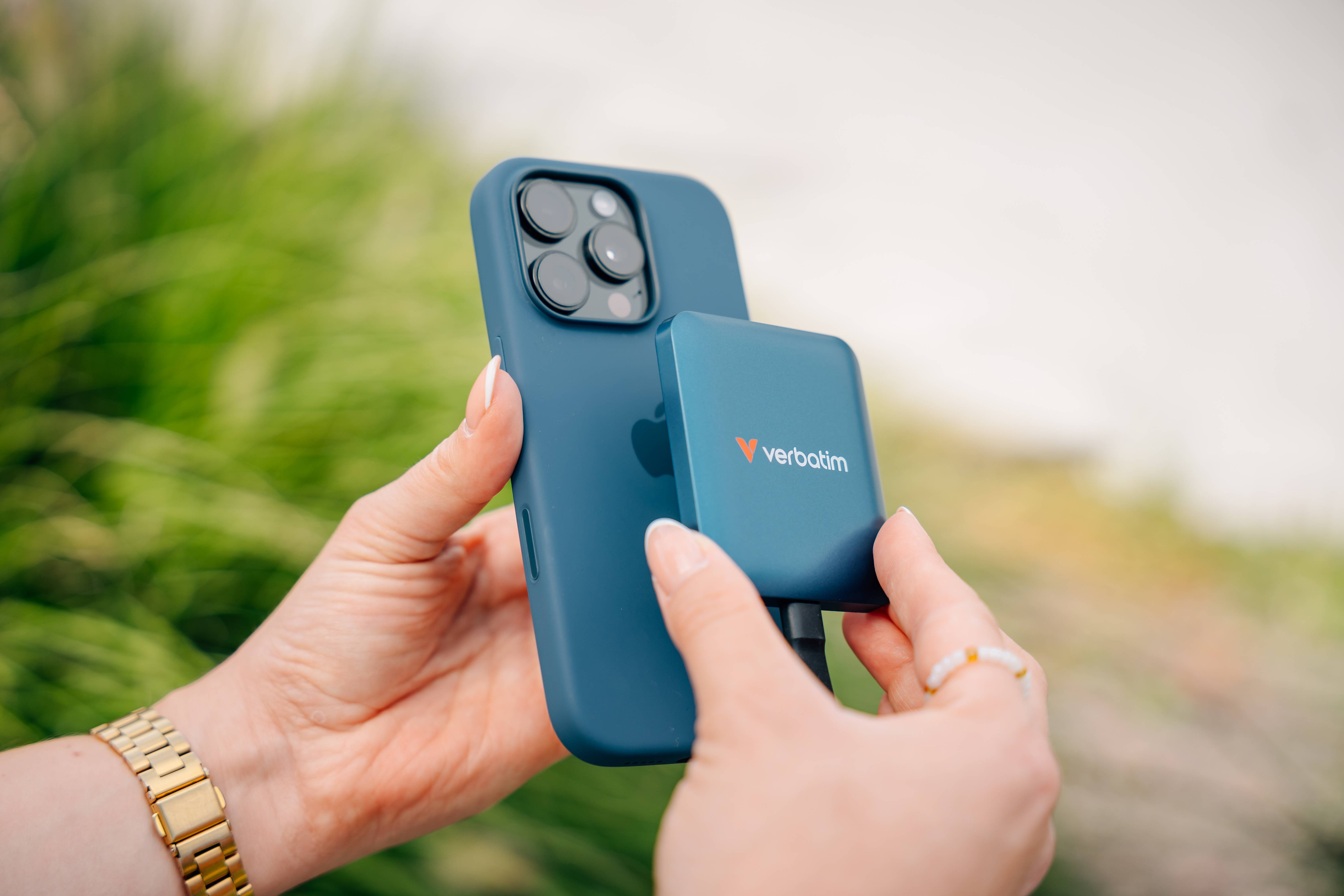 „Eine Hand, die ein Smartphone und eine Verbatim Powerbank hält, mit Grünpflanzen im Hintergrund.