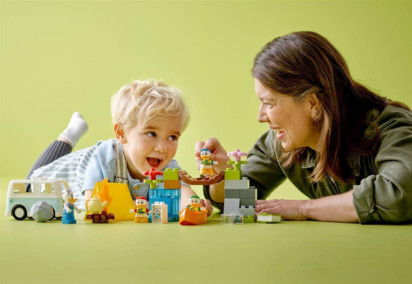 Eine Frau und ein kleines Kind spielen zusammen mit bunten Bausteinen und Spielfiguren auf einer grünen Oberfläche.