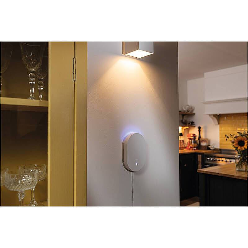 Ein intelligentes Smart-Home-Hub wird an der Wand montiert und sendet ein weiches blaues Licht aus. Es befindet sich in der Nähe eines Schranks mit Glaswaren in einer gemütlichen, modernen Küchenumgebung.