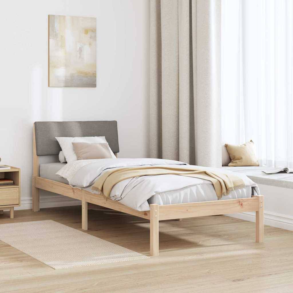 Ein minimalistisches Schlafzimmer mit einem Holzbett, grauem Kopfteil und beiger Bettwäsche. Ein kleiner Nachttisch mit einer Lampe steht links. Weiße Vorhänge hängen am Fenster.