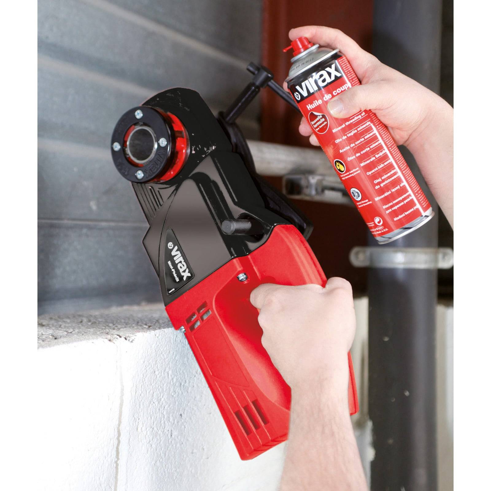 Eine Person verwendet eine rote Sprühdose mit Schmiermittel an einer Gewindemaschine mit der Markenbezeichnung 'virax'. Die Maschine ist auf einem Rohr positioniert.