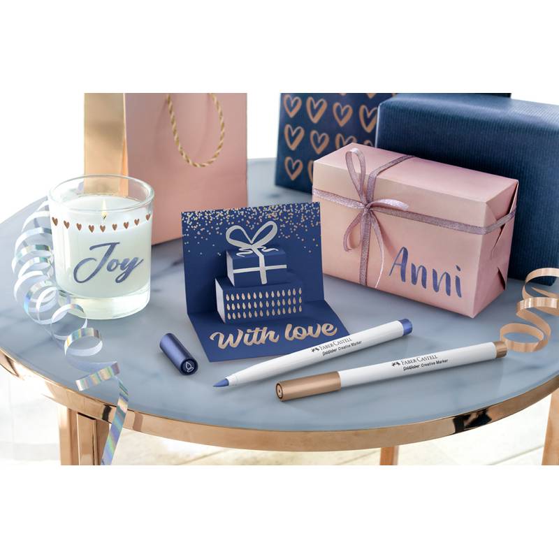 Ein Tisch mit einer Kerze, beschriftet mit 'Freude', eine Karte mit einer Geschenkdesign, Geschenkboxen sowie Stifte, beschriftet mit 'Mit Liebe' und 'Anni'.