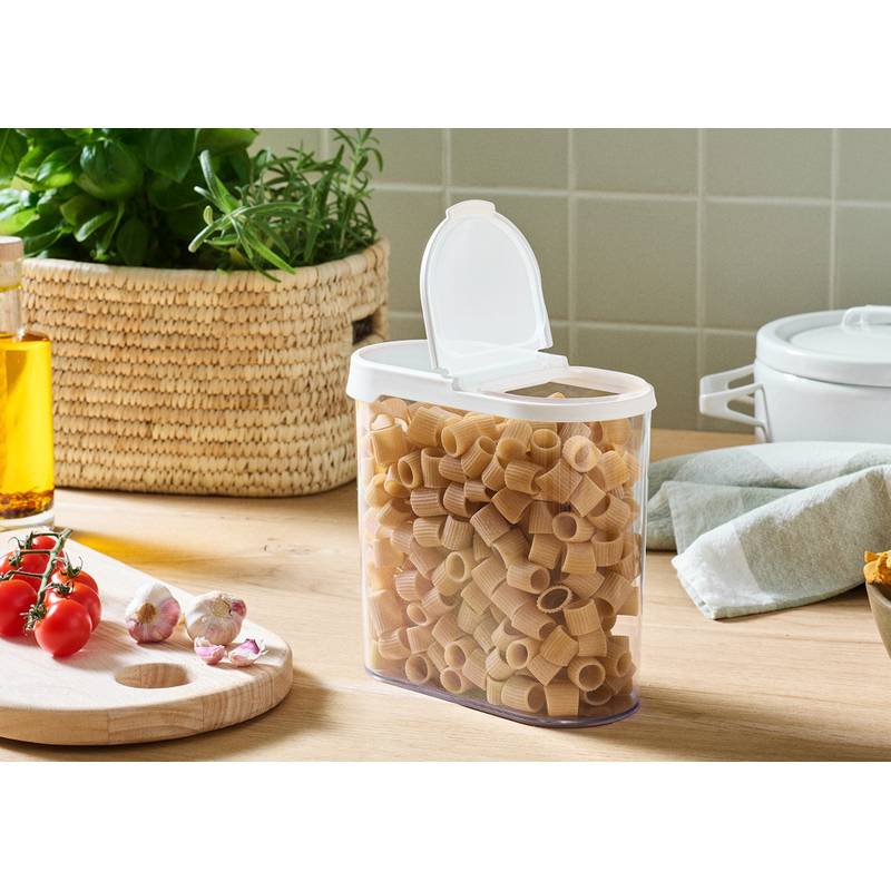 Ein durchsichtiger Behälter gefüllt mit Rigatoni-Nudeln auf einer Küchenarbeitsplatte mit einem Schneidebrett, Tomaten, Knoblauch, Kräutern und einer Ölflasche in der Nähe.
