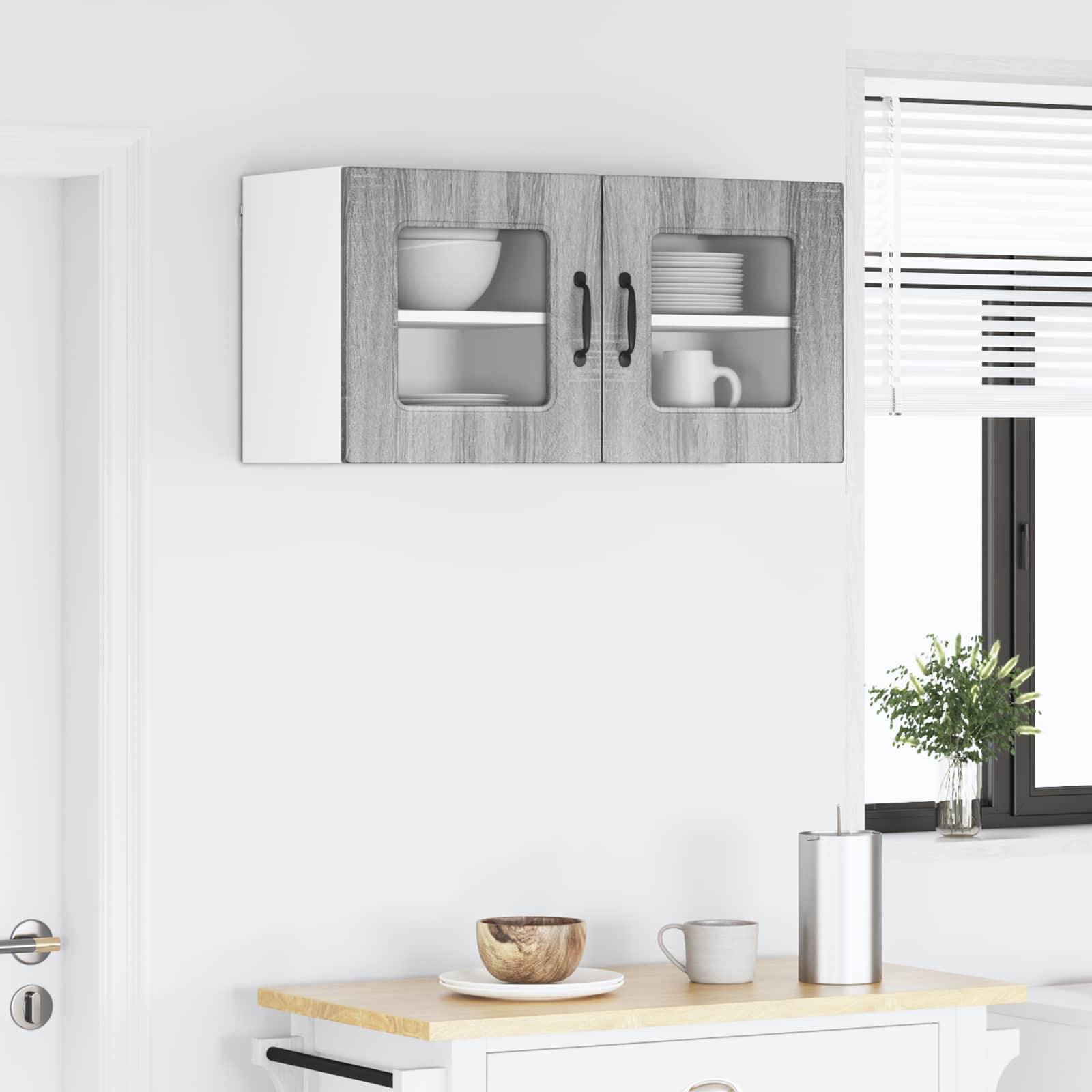 Oberer Küchenschrank mit Glastüren, in dem Geschirr ausgestellt ist. Eine Arbeitsplatte darunter trägt eine Holzschale und einen Metallbecher. Ein nahes Fenster mit einer Topfpflanze.