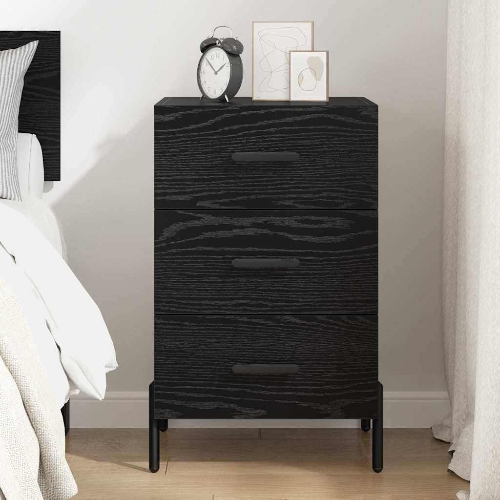 Ein dunkler Holznachttisch mit drei Schubladen steht neben einem Bett. Auf seiner Oberfläche befinden sich ein Wecker und zwei kleine gerahmte Kunstwerke.