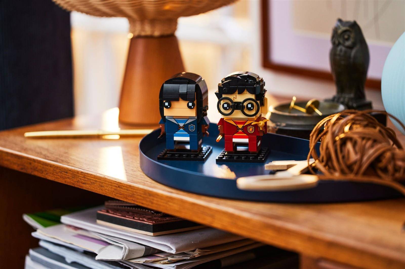 Zwei kleine LEGO-ähnliche Figuren, die einem Mann und einer Frau ähneln, stehen auf einem Tablett auf einem Holztisch mit dekorativen Gegenständen im Hintergrund.