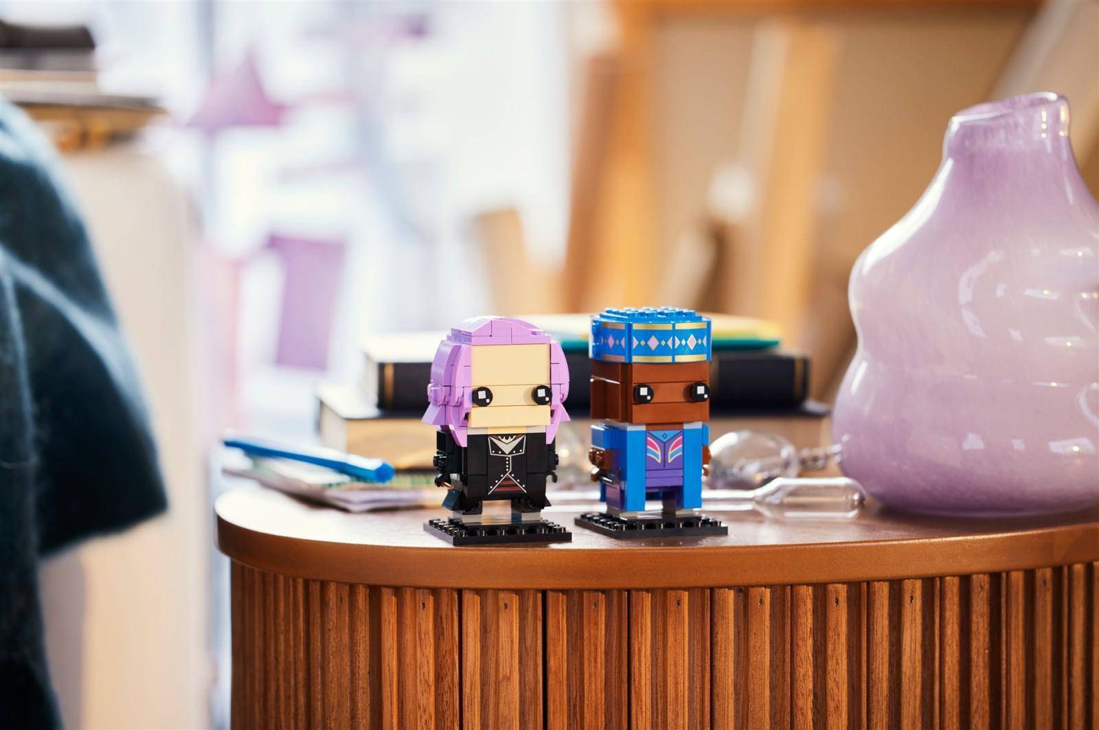 Zwei kleine LEGO-ähnliche Figuren stehen auf einem runden Holztisch neben einer violetten Vase in einem sanft beleuchteten Zimmer.