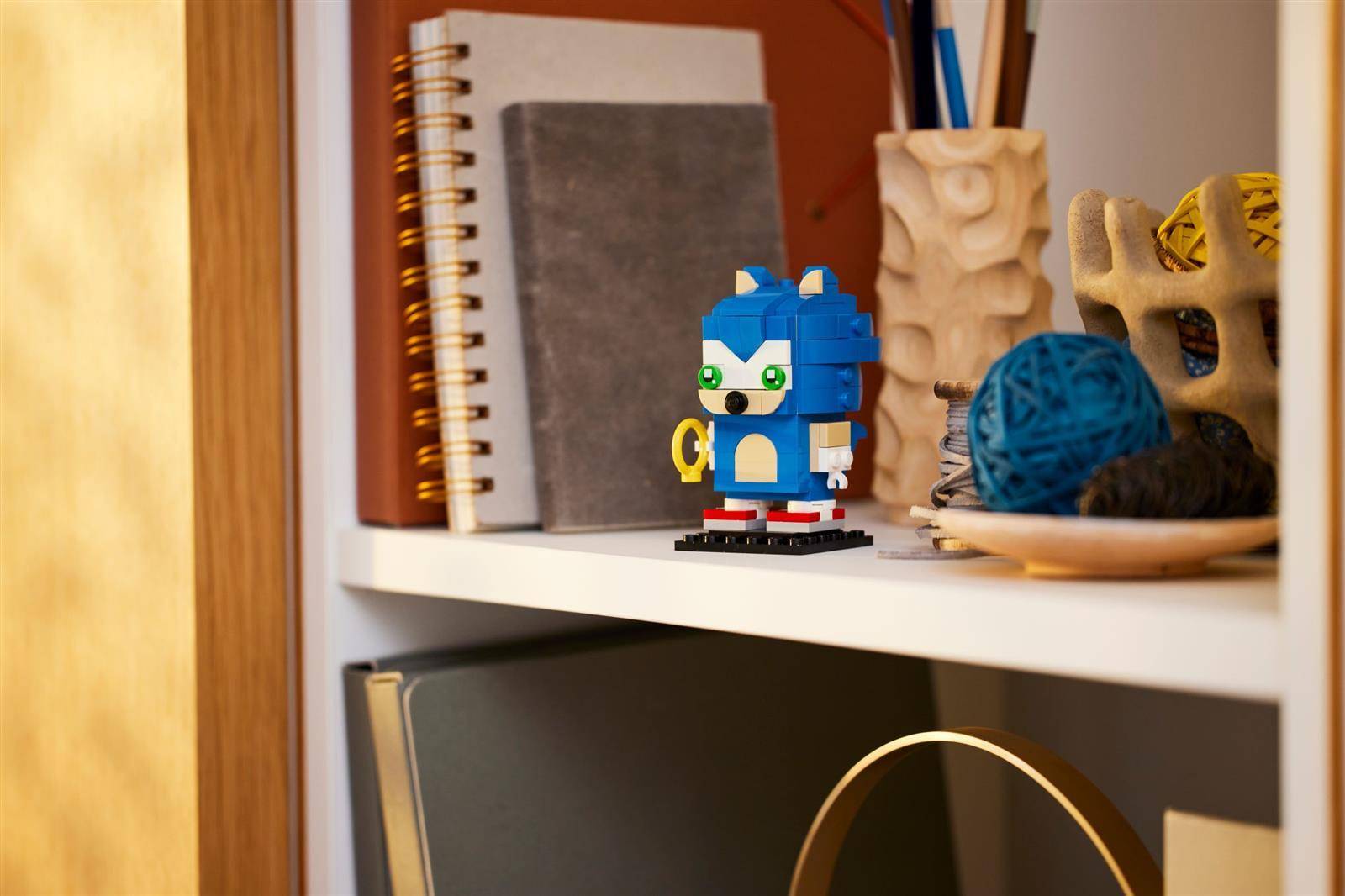 Eine kleine LEGO-Figur von Sonic the Hedgehog sitzt auf einem Regal neben Notizbüchern und Dekorationsobjekten.