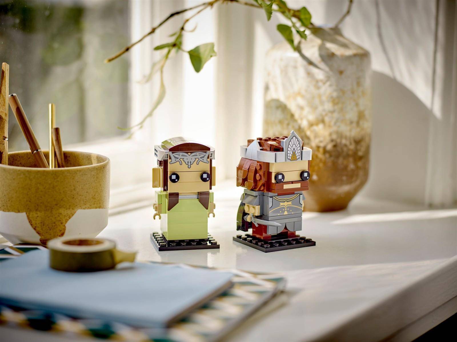 Zwei kleine LEGO-ähnliche Minifiguren-Bauten stehen auf einem sonnigen Schreibtisch neben einem Becher mit Stiften und einer Glasvase.
