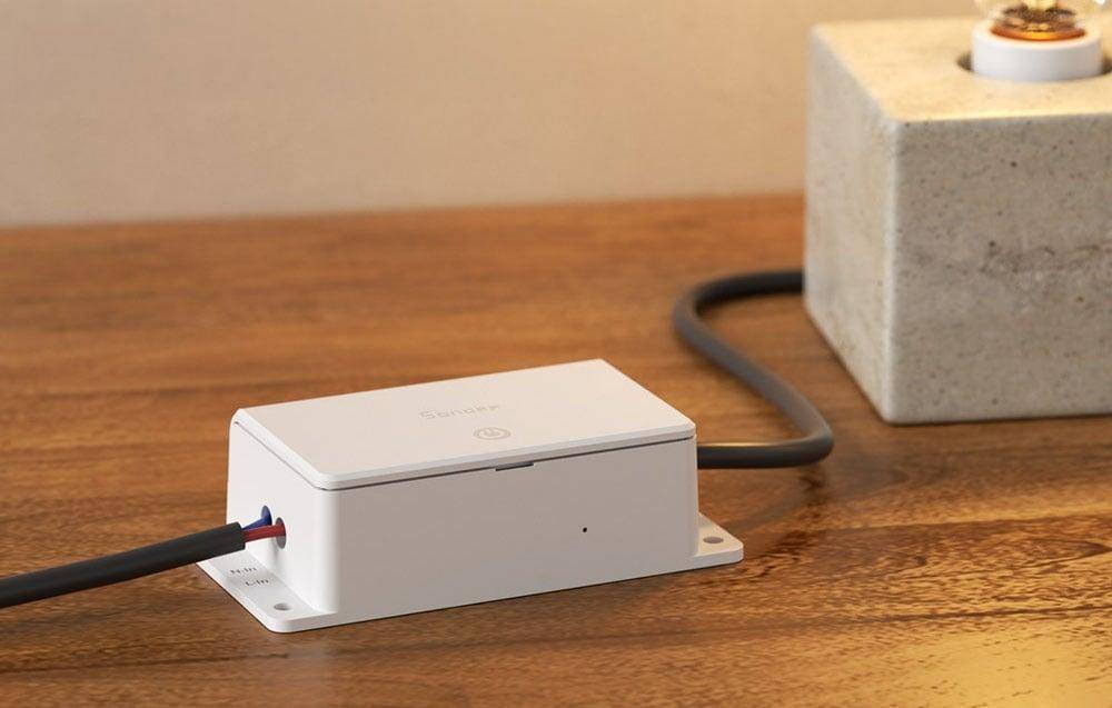 Ein weißer Smart Plug auf einem Holztisch mit angeschlossenem Stromkabel, neben einem steinartigen Objekt. Das Gerät ist mit ‚SwitchBot' beschriftet.