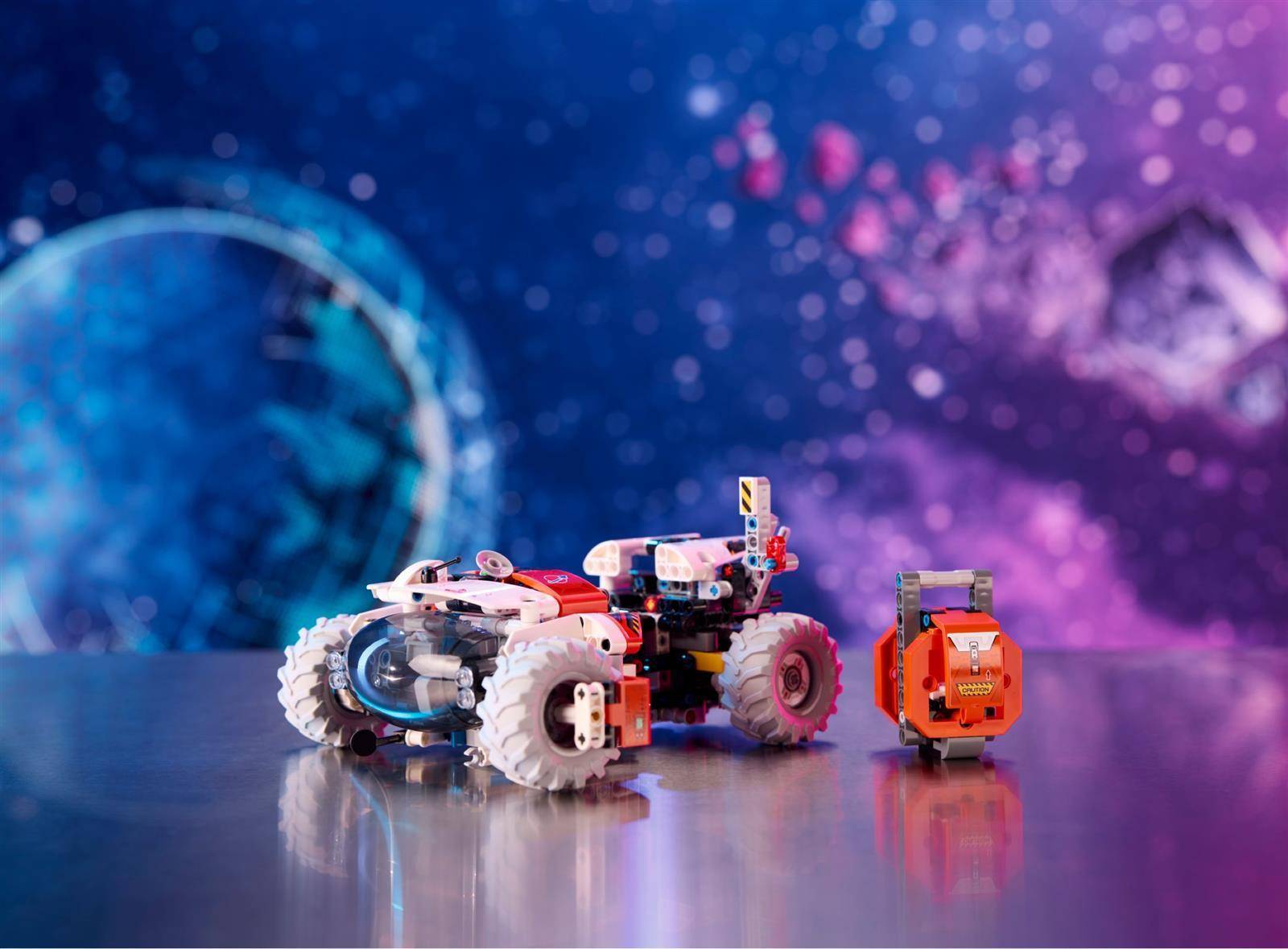 Zwei kleine Spielzeugfahrzeuge, ein größeres Roboter-Auto mit weißen Rädern und ein kleinerer orangefarbener Wagen, stehen auf einer reflektierenden Oberfläche vor einem blau-violetten abstrakten Hintergrund.