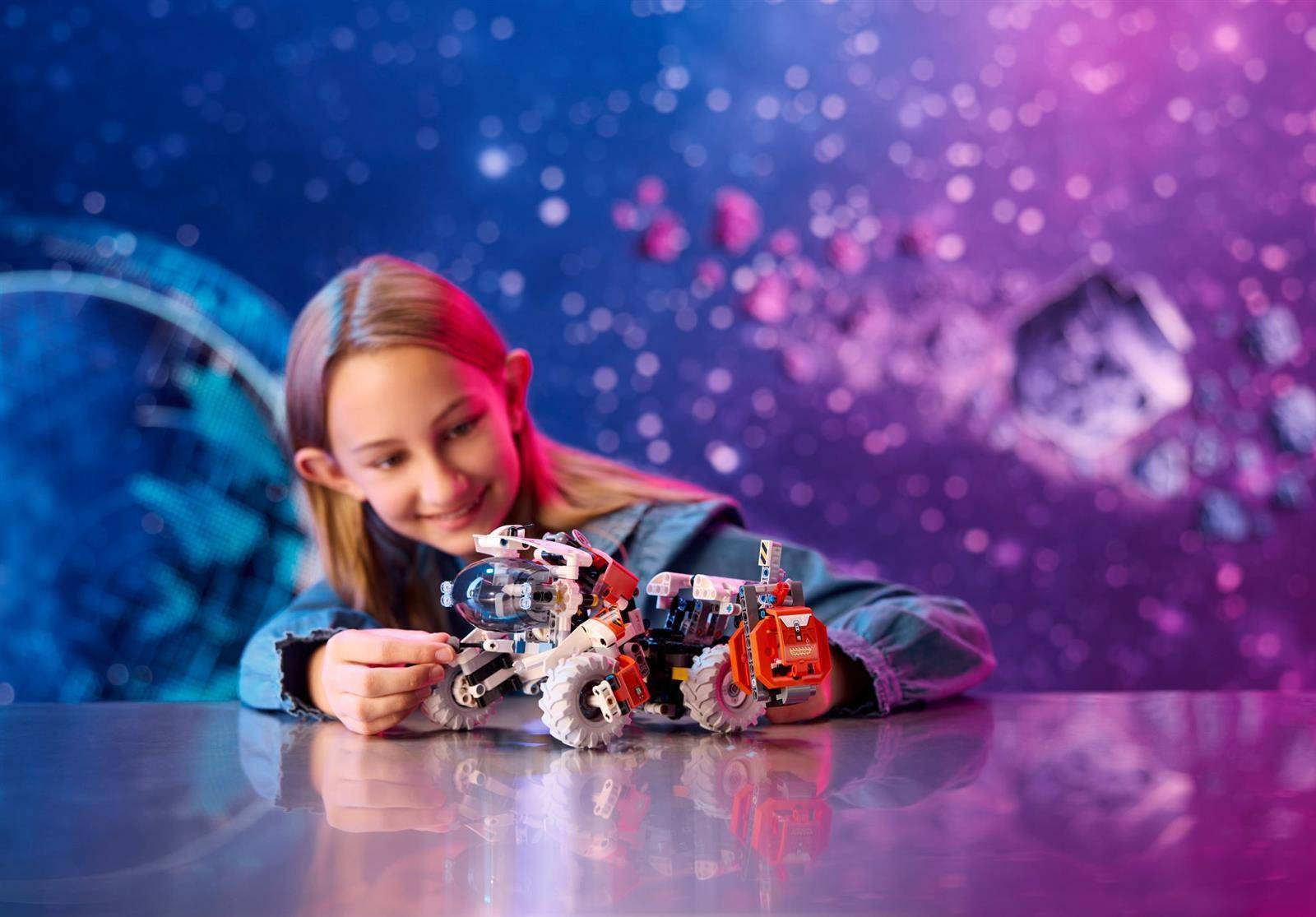 Ein lächelndes Mädchen lehnt sich über einen Tisch und stellt ein kleines Roboterfahrzeug mit einem Greifarm vor einem blau-violetten, technologiethematischen Hintergrund ein.