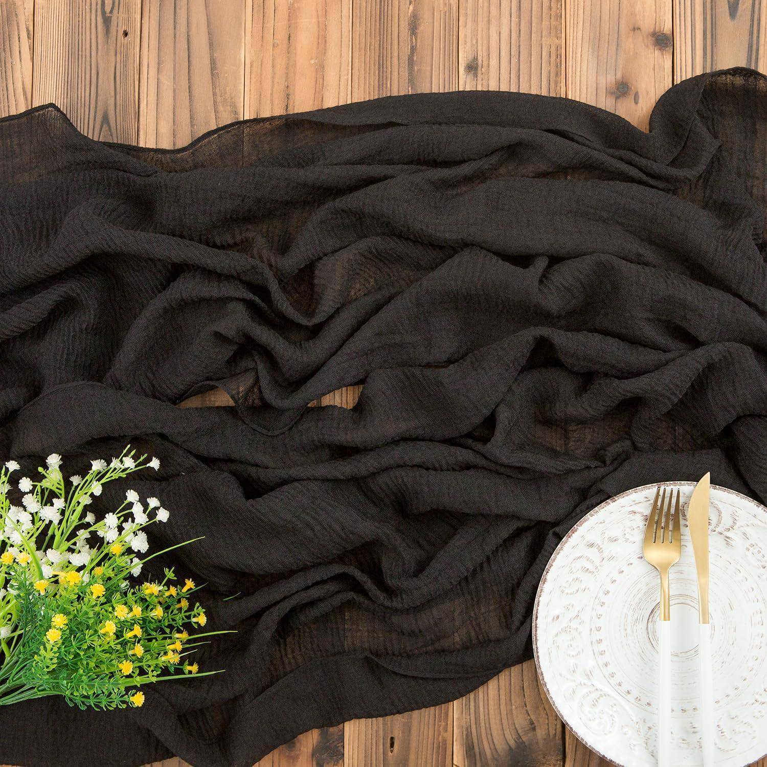 Ein schwarzer Stoff ist elegant auf einer Holzoberfläche drapiert, begleitet von einem Blumenstrauß aus kleinen weißen und gelben Blüten sowie einem weißen Teller mit einer goldenen Gabel und einem Messer.