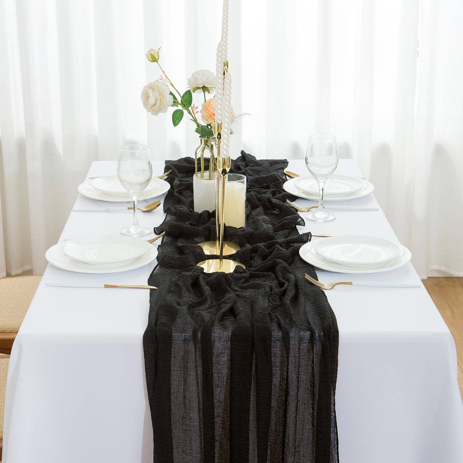 Ein weißer Tisch mit einem schwarzen Läufer und vier Geddecken, jeweils mit einem Teller, Besteck und Weinglas. Ein Mittelstück mit Blumen und Kerzen.