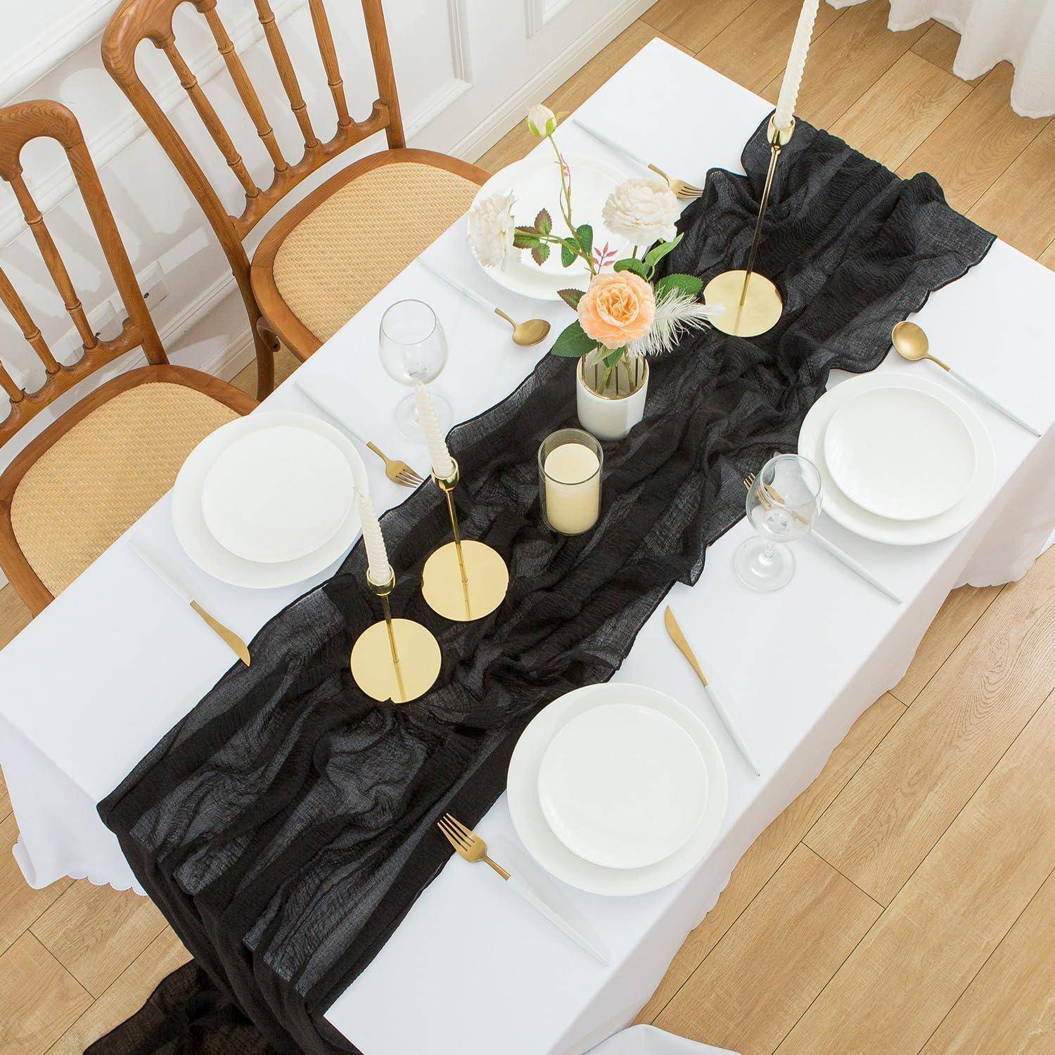 Tisch elegant gedeckt mit weißen Tellern, goldenen Bestecken und Gläsern auf einer weißen Tischdecke. Ein schwarzer Läufer, Blumen und Kerzen schmücken die Mitte.