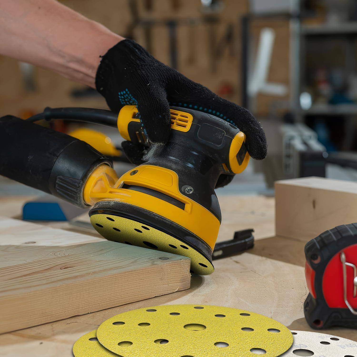 Eine Person mit Handschuh verwendet einen gelb-schwarzen elektrischen Schleifer auf einem Holzbrett, wobei Schleifscheiben und ein Maßband in der Nähe liegen.