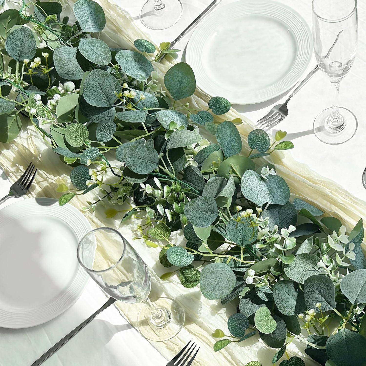 Ein für ein formelles Essen gedeckter Tisch mit weißen Tellern, leeren Gläsern und einem Mittelstück aus üppigem Grün und kleinen weißen Blumen auf einer hellen Tischdecke.
