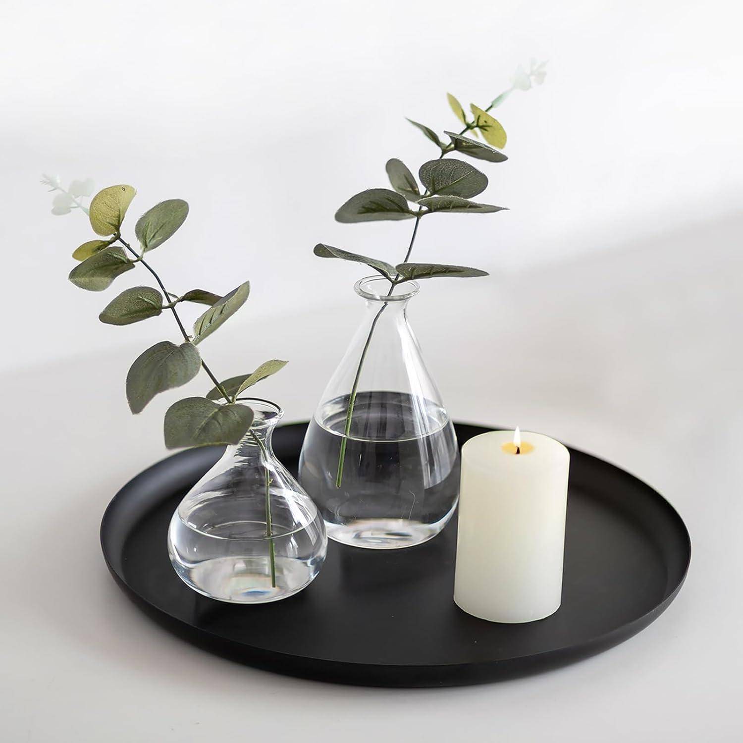 Ein schwarzes Tablett trägt zwei klare Glasvasen mit grünen, blattreichen Stängeln und eine weiße Säunenkerze, wodurch eine minimalistische Dekoration entsteht.