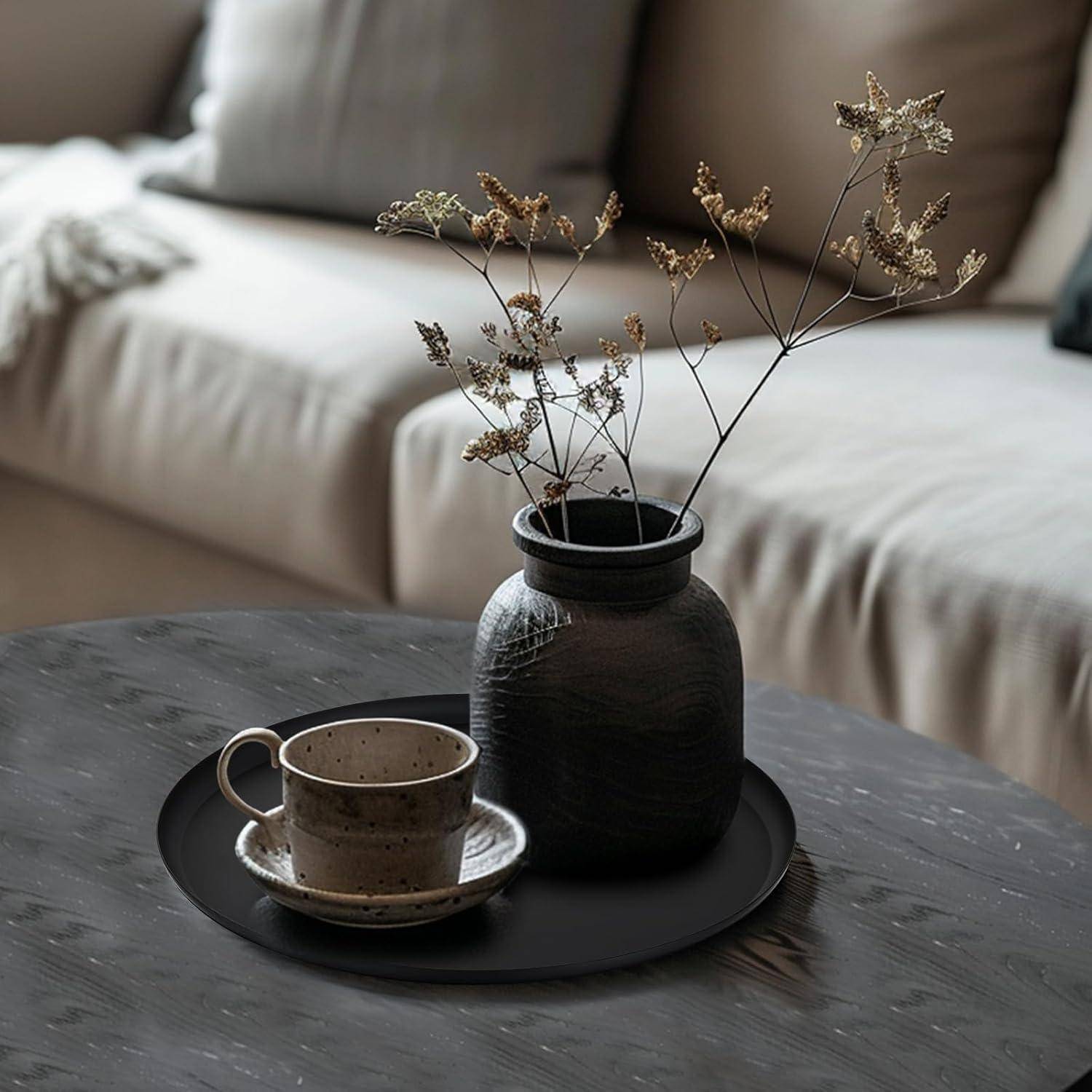 Eine schwarze Vase mit getrockneten Blumen und einer Keramik-Teetasse auf einem dunklen runden Tablett, das auf einem Holzkaffeetisch in einem gemütlichen Wohnzimmer steht.