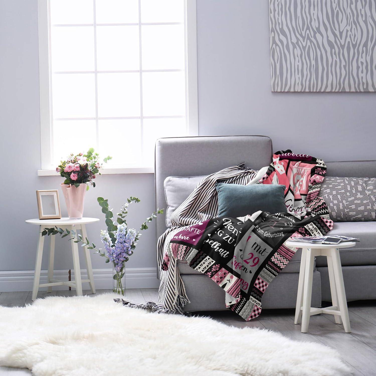 Ein gemütliches Wohnzimmer mit einem grauen Sofa, das mit einer bunten Decke bedeckt ist, einem kleinen Beistelltisch mit einer Vase voller Blumen und einem großen Fenster, das natürliches Licht hereinlässt.