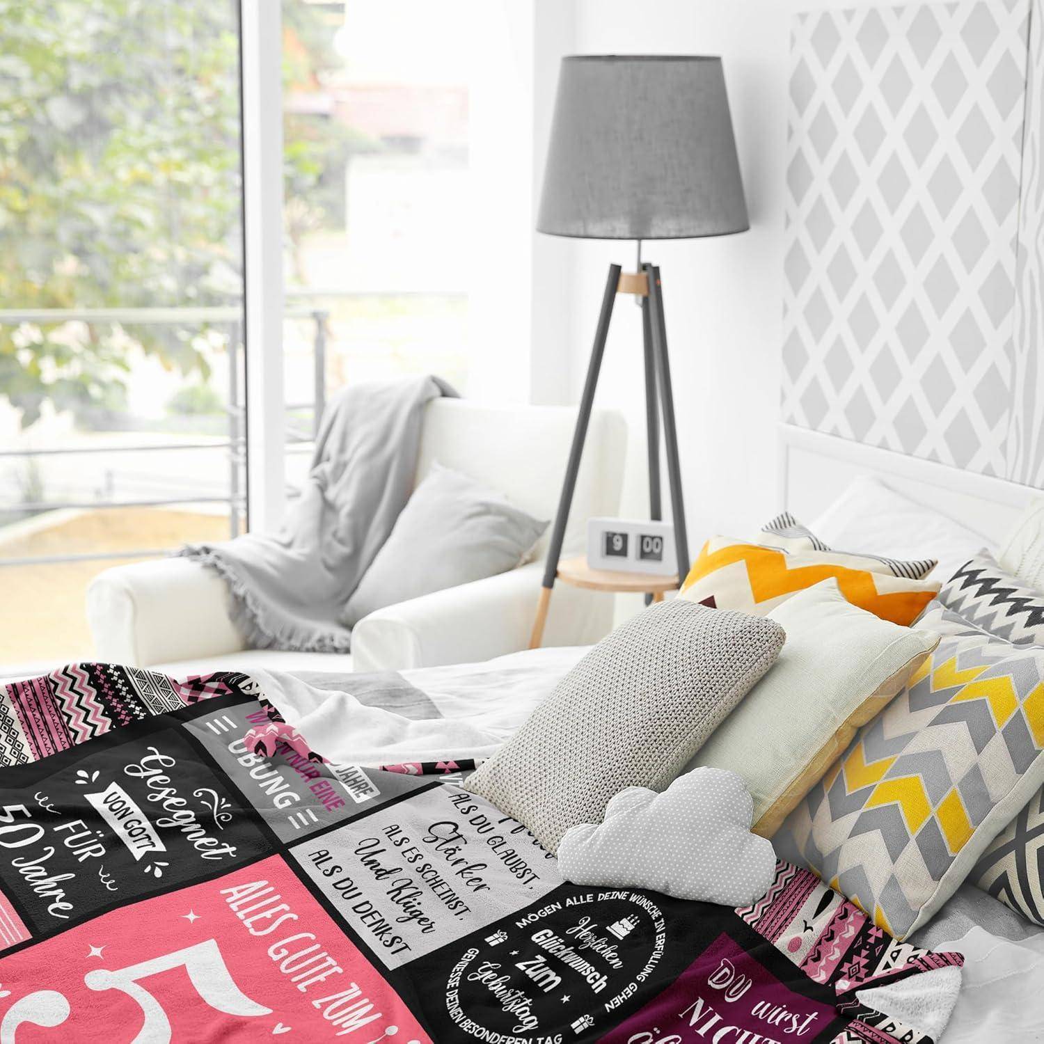 Ein ordentlich gemachtes Bett mit gemusterten Decken, die inspirierende Zitate tragen, ist umgeben von bunten Kissen und einer grauen Lampe in einem hellen Raum.