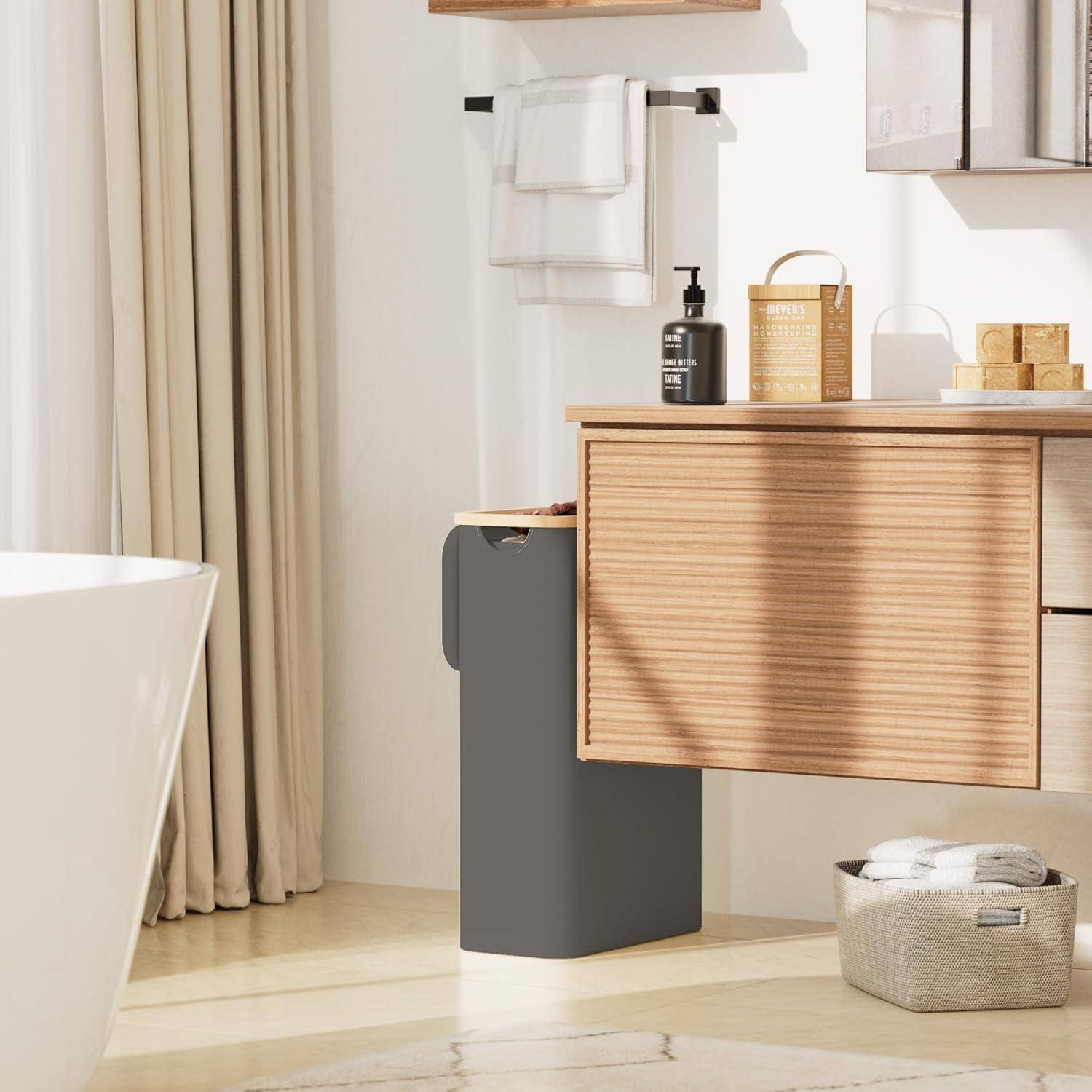 Modernes Badezimmer mit Waschtisch aus Holz, schwarzem Seifenspender, weißen Handtüchern, grauem Mülleimer und geflochtener Korb auf dem Boden; sonnendurchfluteter Raum.