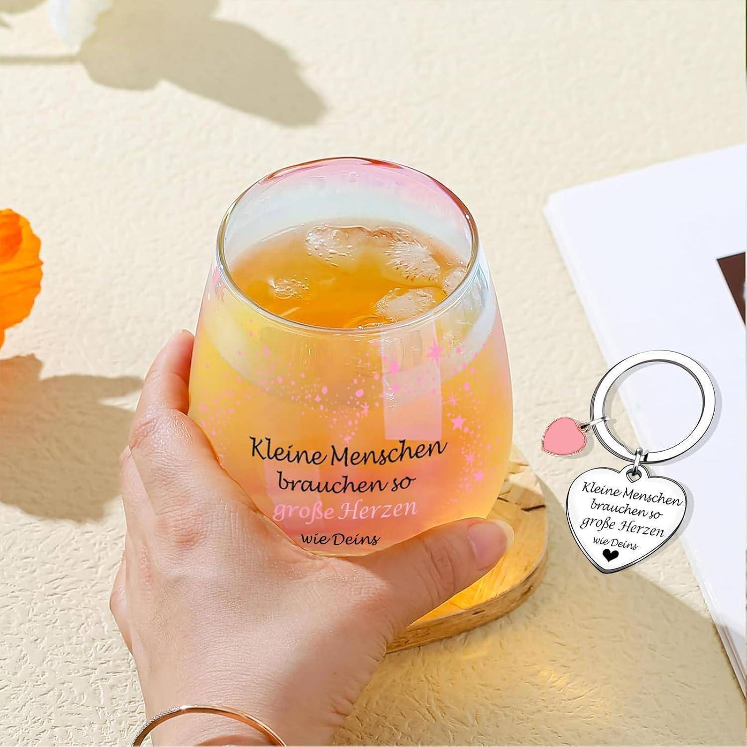 Eine Hand hält ein Glas mit orangefarbener Flüssigkeit. Das Glas trägt den Text: 'Kleine Menschen brauchen so große Herzen wie Deins.' Ein Schlüsselanhänger mit einem ähnlichen Text befindet sich daneben.