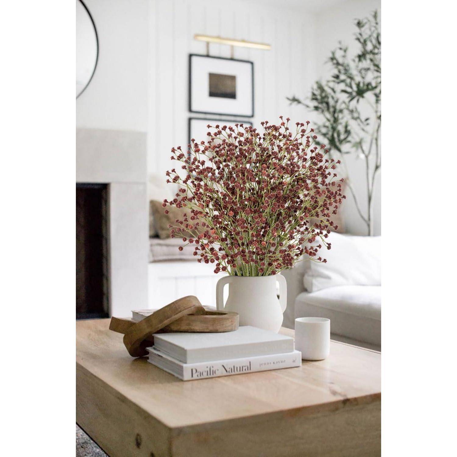 Eine weiße Keramikvase mit roten Blumen auf einem Holzkaffeetisch, umgeben von Büchern und einer Kerze, in einem modernen Wohnzimmer.
