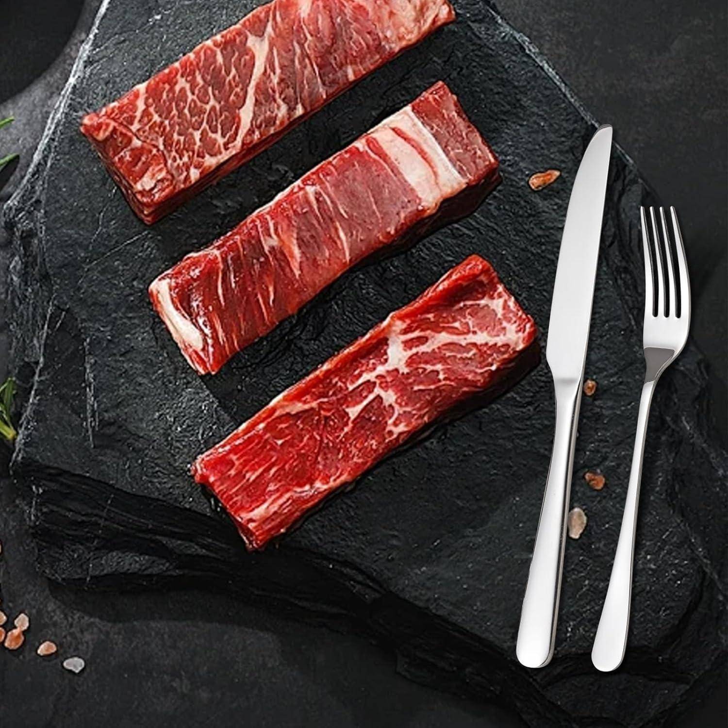 Drei rohe Steakstreifen liegen mit einem Messer und einer Gabel auf einer dunklen Schieferoberfläche, bereit zum Kochen oder Servieren.