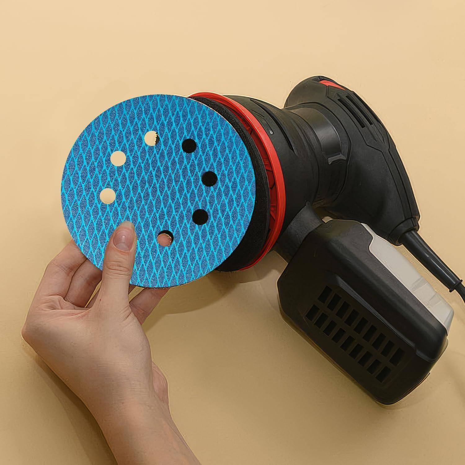 Eine Hand hält eine blaue Schleifscheibe mit sieben Löchern in der Nähe eines elektrischen Schleifgeräts. Das Schleifgerät ist schwarz mit roter Einfassung und einem Netzkabel.