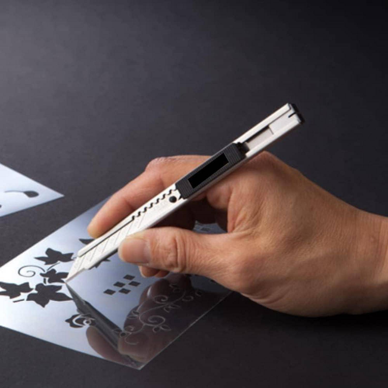 Eine Hand verwendet ein Bastelmesser, um Formen aus einem dekorativen Schablonenmuster auf dunklem Papier auszuschneiden, wobei der Fokus auf Präzision und Details liegt.