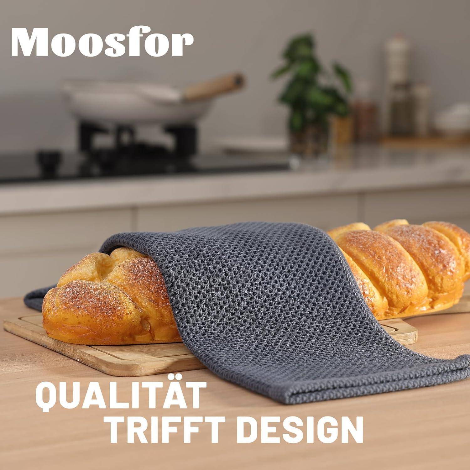 Ein Laib Brot auf einem Schneidebrett, das mit einem grauen strukturierten Tuch bedeckt ist, wobei der Text ‚Qualität trifft Design' und ‚Moosfor' sichtbar sind.