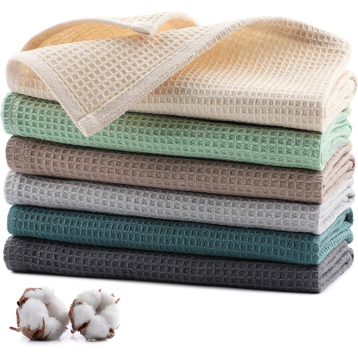 Ein Stapel von farbigen Waffelpiquee-Handtüchern in Beige, Grün, Grau und Petrol, mit zwei Baumwollblüten davor.