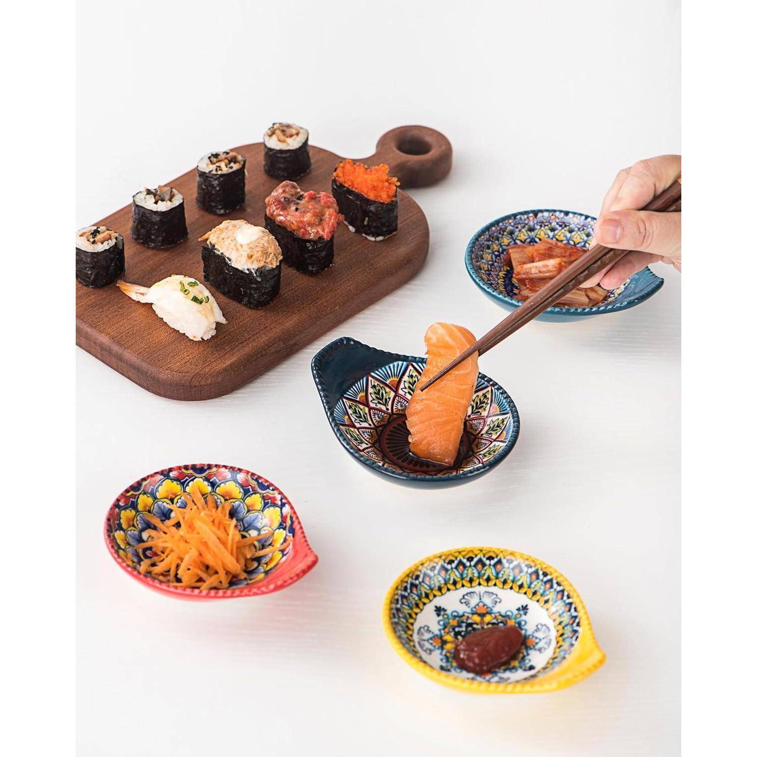 Eine Hand benutzt Essstäbchen, um ein Lachssushi in Sojasauce zu tauchen. Verschiedene Sushistücke und bunte Schälchen mit Beilagen befinden sich auf einer weißen Oberfläche.