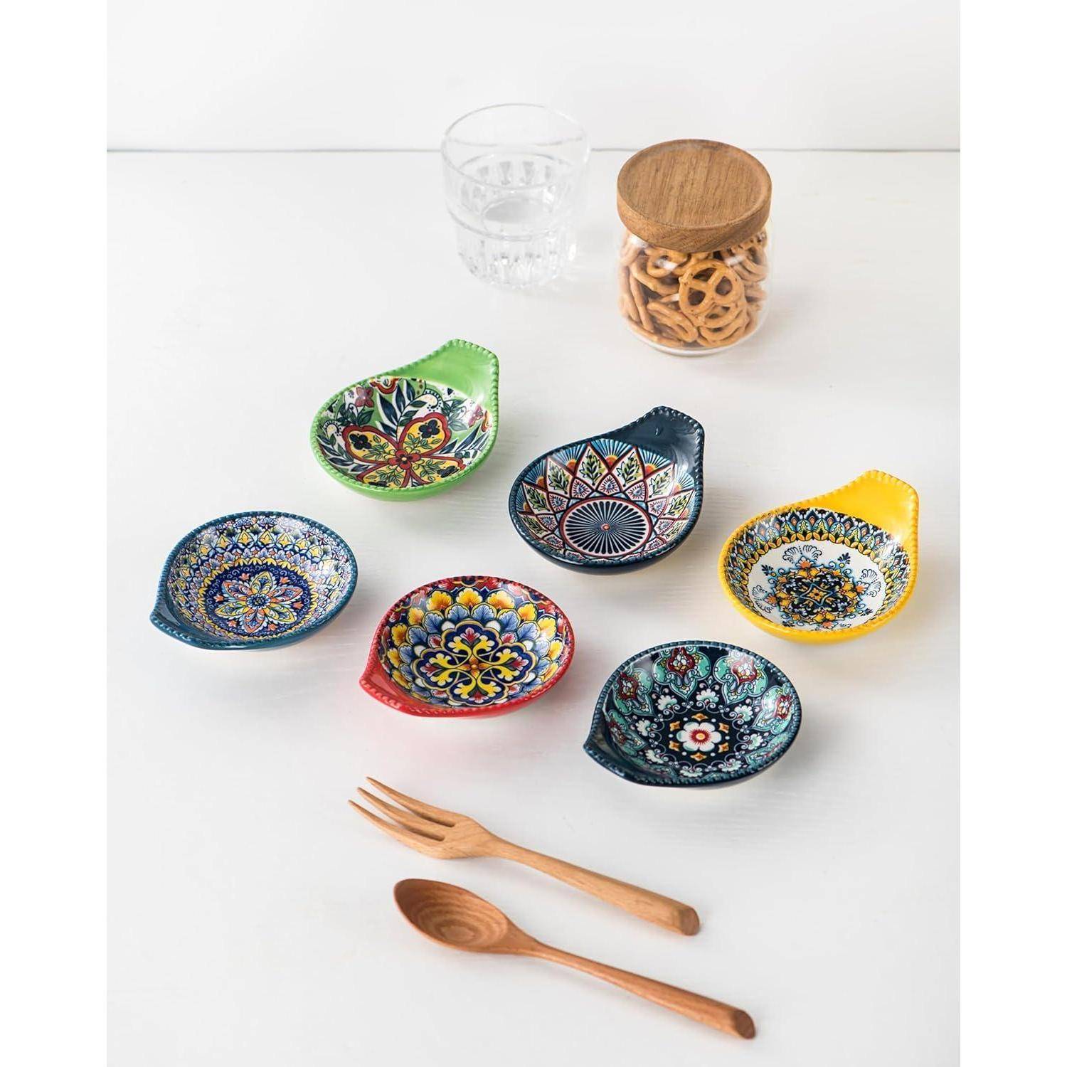 Bunte Keramik-Untertassen, die auf einem weißen Tisch angeordnet sind, mit einem Holzgefäß voller Snacks, einem Wasserglas und Holzutensilien in der Nähe.