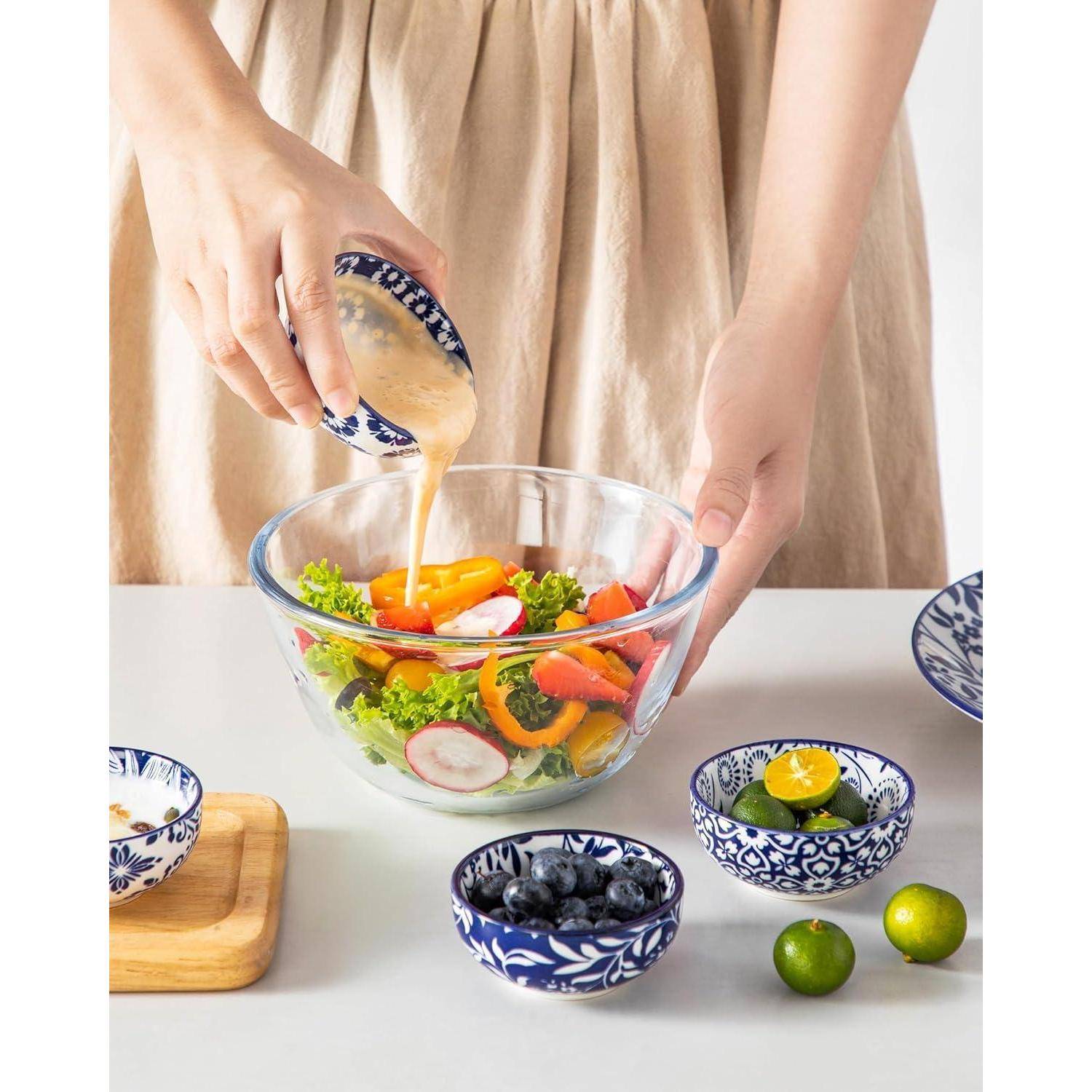 Eine Person gießt Dressing in eine Salatschüssel mit Gemüse, darunter Salat, Radieschen und Paprika. Blaue gemusterte Schüsseln mit Obst stehen daneben.