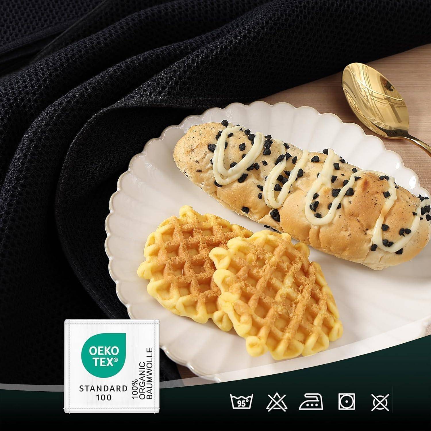 Eine Platte mit einem Croissant, das mit Zuckerguss und Samen verziert ist, begleitet von zwei Waffelstücken. Ein goldfarbener Löffel liegt in der Nähe. Oeko-Tex-Etikett ist sichtbar.
