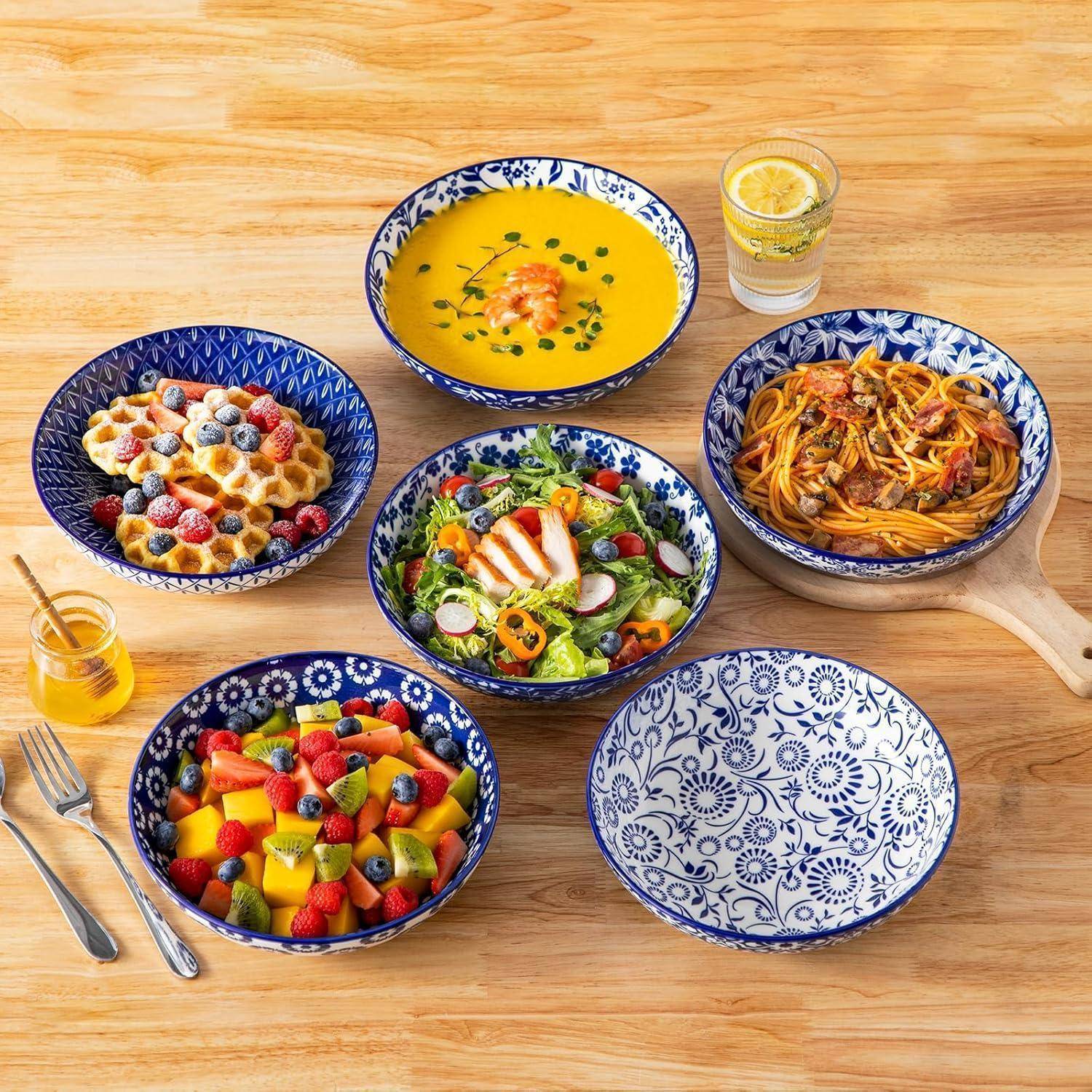 Sieben blau gemusterte Schüsseln auf einem Holztisch, die verschiedene Speisen enthalten: Suppe, Salat, Obstsalat, Pasta, Waffeln mit Beeren und eine leere Schüssel. Ein Glas Limonade und ein kleines Honigglas befinden sich ebenfalls darauf.