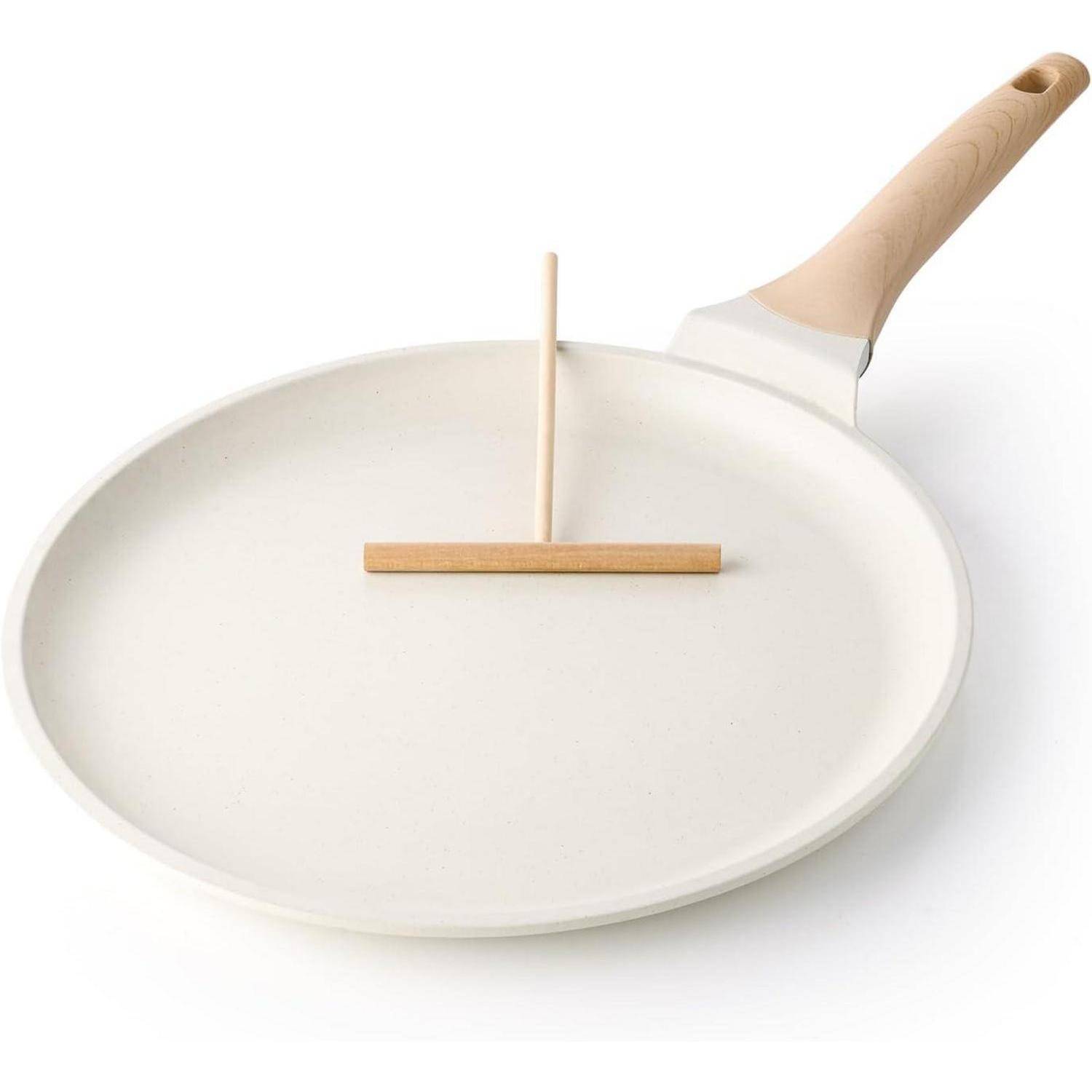 Eine weiße Crêpepfanne mit einem Holzgriff und einem Holzspatel, der auf ihrer Oberfläche liegt, und die zum Zubereiten von Crêpes konzipiert ist.